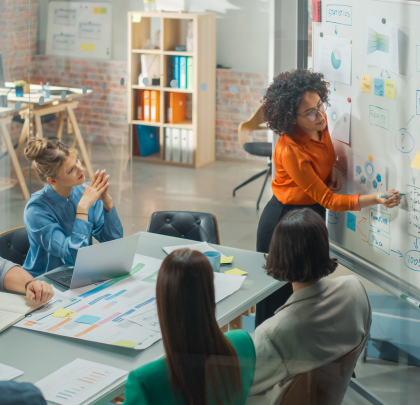 A group of colleagues gather around a table while one of them illustrates ideas on a whiteboard